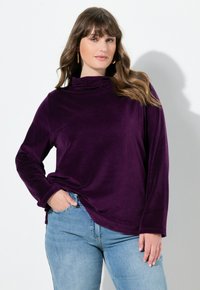 Lila Langarmshirt aus weichem Stoff mit hohem Kragen und lockerer Passform. Kombiniert mit hellblauen Jeans, die den lässigen Stil betonen.