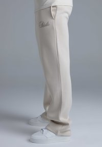 Light beige sweatpants with a loose fit, featuring side pockets and an embroidered logo. Paired with white platform sneakers.
