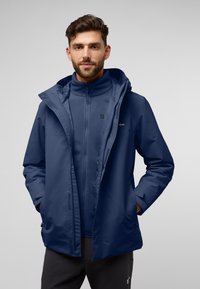 Giacca impermeabile blu navy con cappuccio, zip frontale e polsini elasticizzati, indossata sopra un pile coordinato, con una grafica minimale.