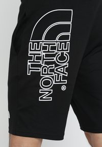 Pantaloni corti neri in cotone con una texture liscia. Presentano un grande logo bianco stampato sul lato e tasche laterali per riporre oggetti.