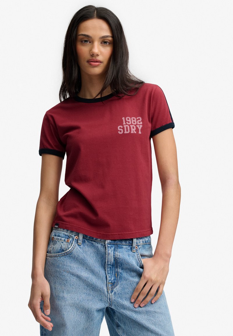 Donna con capelli lunghi e scuri che indossa una maglietta rossa con bordi neri e la scritta "1982 SDRY", abbinata a jeans in denim azzurro chiaro.