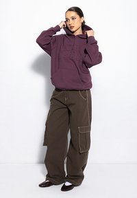Fioletowa bluza z kapturem z wytłoczonym tekstem, w połączeniu z luźnymi oliwkowymi spodniami cargo z kieszeniami i kontrastującym szwem. Brązowe buty dopełniają wygląd.