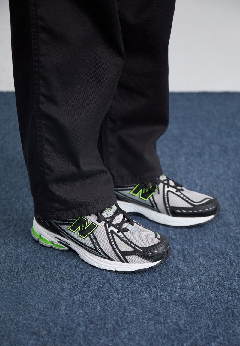 Personne portant un pantalon noir et des chaussures de course New Balance blanches, noires et vertes debout sur un tapis bleu.