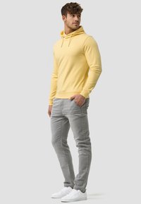 Gul hættetrøje med ribbede manchetter og kant, parret med lysegrå slim-fit jeans og hvide sneakers. Minimalistisk design, ingen mønstre.
