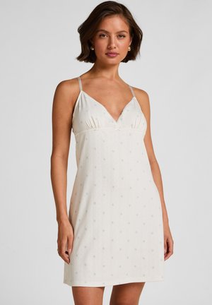 Jonge vrouw met kort bruin haar draagt een mouwloze witte minijurk met dunne bandjes en een subtiel klein bloemenpatroon, poserend tegen een effen achtergrond.