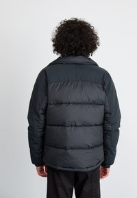 Sort pufferjakke med horisontale quiltede sektioner, en fold-down krave og lange ærmer. Materialet fremstår blankt og polstret for varme.