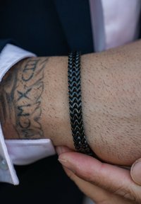 Black braided bracelet on a wrist, featuring a textured design. Skin is visible beneath the bracelet, with a tattoo partially showing.