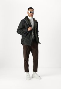 Dark green waterproof jacket with pockets, worn over a white turtleneck. Brown corduroy pants and white sneakers complete the outfit. Silver sunglasses worn.