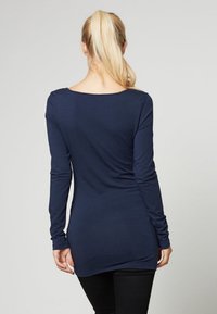 Granatowa bluzka z długim rękawem, dekoltem w serek, dopasowanym krojem i miękką fakturą. Nosi z czarnymi spodniami, prezentując widok z tyłu.