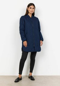 Marinblå quiltad jacka med hög krage, frontdragkedja och långa ärmar. Bärs över svarta åtsittande byxor och glansiga loafers.