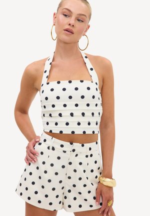 Kobieta ubrana w biały crop top typu halter i pasujące szorty w czarne kropki, złote kolczyki koła, złotą bransoletkę oraz pierścionek w kształcie słońca.