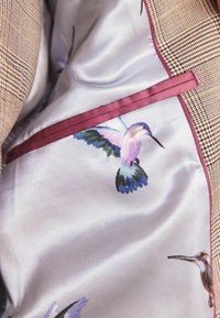 Doublure de veste en soie avec des imprimés colorés de colibris à l'intérieur d'un blazer marron à carreaux avec un liseré bordeaux autour de la poche intérieure.
