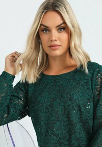 Mörkgrön spetsblus med blommönster, långa ärmar med genomskinliga detaljer och spridda paljetter för extra textur och detalj.