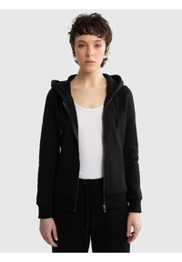 Tânăr cu păr scurt și creț, purtând un hanorac negru cu fermoar peste un maiou alb și pantaloni negri cu șnur, stând într-un fundal simplu.