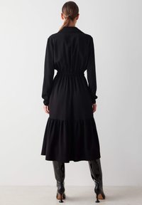 Nainen, jolla on ruskea ponihäntä, käyttää pitkähihainteista mustaa midi-mekkoa, jossa on kerrostettu helma ja gathered-vyötärö, yhdistettynä mustiin kiiltonahkaisiin polvipitkiin saappaisiin.