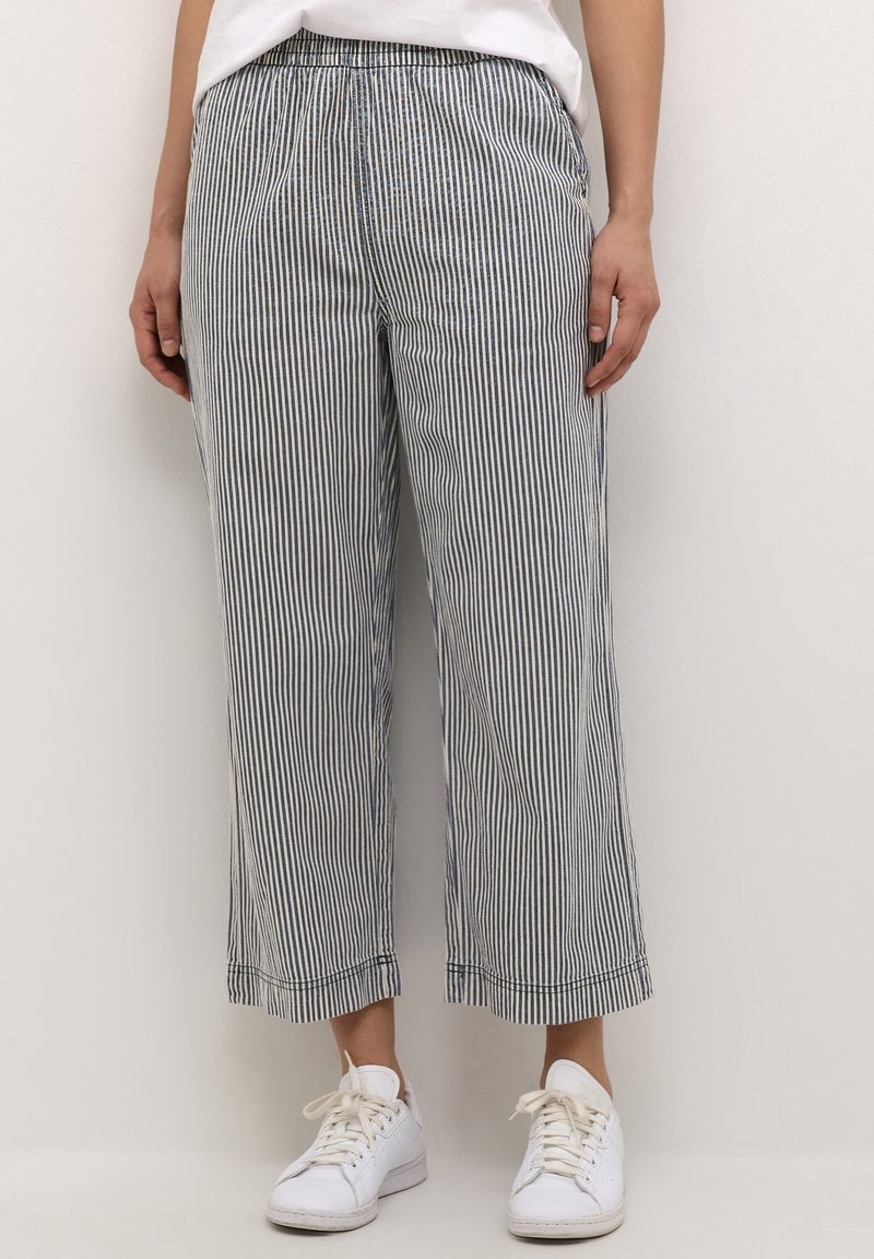 Personne portant un pantalon taille haute, ample, longueur cheville avec de fines rayures verticales bleu marine et blanc et des baskets blanches.