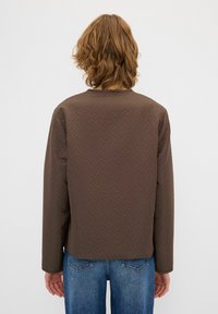 Giacca trapuntata marrone con un motivo a spina di pesce, maniche lunghe, colletto rotondo e vestibilità comoda, abbinata a jeans blu.