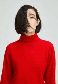 Roter Strickrollkragenpullover mit weicher Textur, der eine taillierte Passform und überschnittene Schultern hat, teilweise von Haaren verdeckt.
