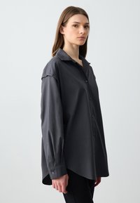 Chemise grise à manches longues, en tissu lisse, avec un col, des fentes sur les côtés et des détails discrets aux poignets. Ajustement tailleur.