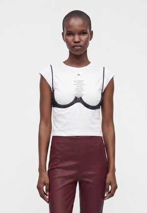 Γυναίκα με κοντά μαλλιά φορά λευκό κομμένο t-shirt με μαύρο σουτιέν από πάνω και ψηλόμεσο σκουροκόκκινο παντελόνι, στέκεται μπροστά σε λευκό φόντο.