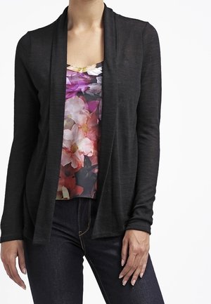 Femme portant un cardigan noir ouvert sur un haut à imprimé floral et un jean foncé, debout avec une main sur la cuisse.