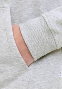 Ręka częściowo włożona w przednią kieszeń jasnoszarego swetra z prążkowanym mankietem i widocznymi detalami stębnowania.