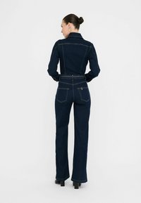 Jumpsuit in denim scuro con un corpetto aderente, maniche lunghe e pantaloni a gamba larga. Presenta un punto vita con cintura e tasche posteriori con cuciture a vista.