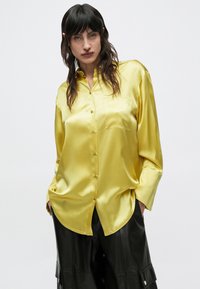 Άτομο με μακριά σκούρα μαλλιά που φοράει ανοιχτόχρωμο, λαμπερό κίτρινο πουκάμισο με κουμπιά και μαύρο, φαρδύ δερμάτινο παντελόνι, στέκεται μπροστά σε απλό φόντο.