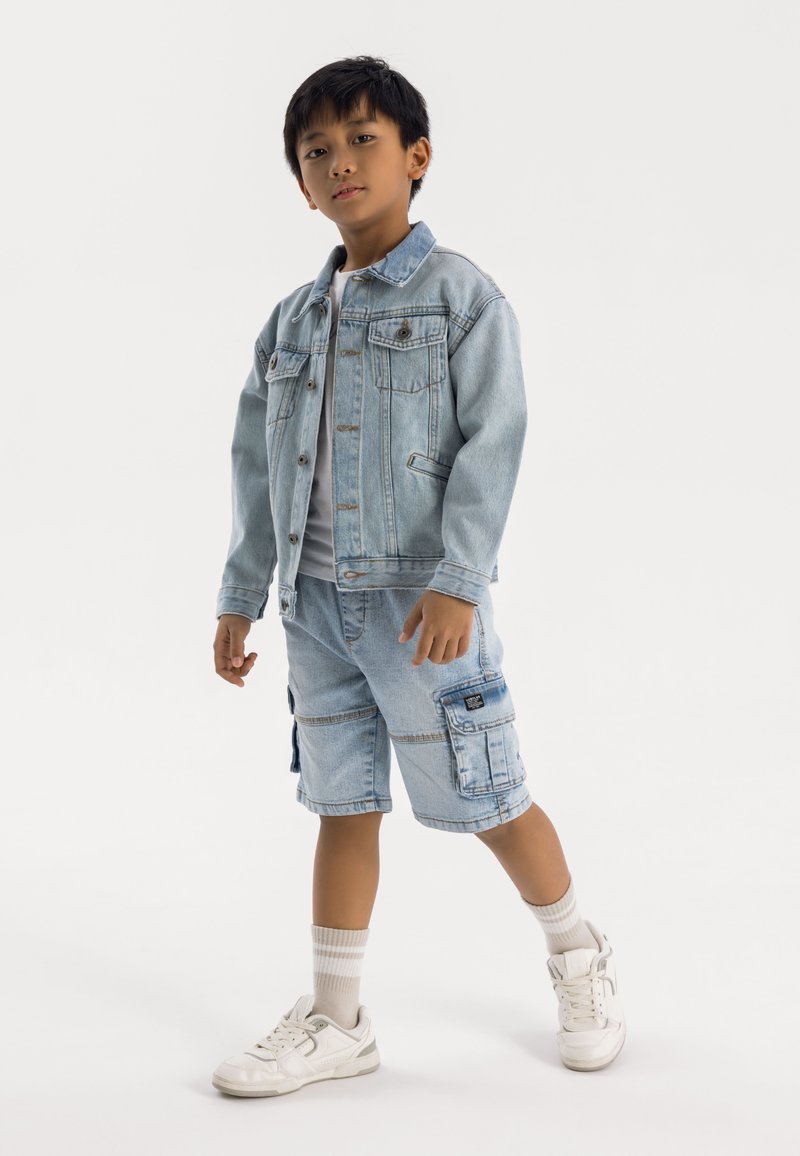 Denim jakna, svetlo modra, z gumbnim zapiranjem in žepom na prsih. Ujemajoče svetlo modre cargo kratke hlače z stranskimi žepi. Bele superge in sivi nogavice.