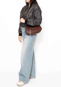 Borsa a tracolla in pelle marrone con una finitura texturizzata, forma slanciata, dettagli con zip e tracolla staccabile, indossata con una giacca bomber nera e jeans chiari.