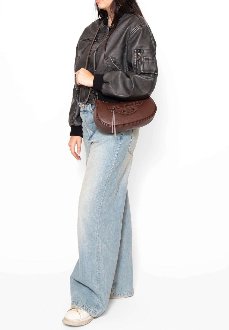 Borsa a tracolla in pelle marrone con una finitura texturizzata, forma slanciata, dettagli con zip e tracolla staccabile, indossata con una giacca bomber nera e jeans chiari.