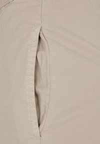 Gros plan d'une poche de pantalon beige avec ouverture verticale et coutures visibles sur tissu en coton.