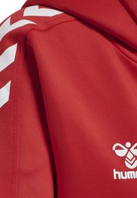 Czerwona tkanina z białymi ukośnymi paskami na krawędzi i białym logo Hummel w rogu, pokazująca część ubrania.