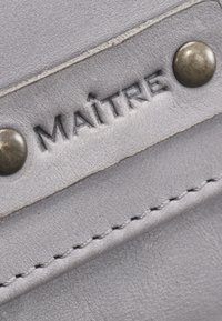 Graues Lederstück mit geprägtem "MAÎTRE", versehen mit Messingnieten und feiner Naht am Rand, das Textur und Handwerkskunst präsentiert.