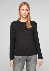 Pull en maille noir avec motif texturé, col rond et manches longues. Associé à un jean gris, souligné par un design minimaliste.