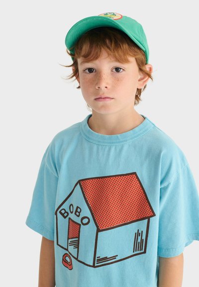 Jeune garçon avec des taches de rousseur portant une casquette verte et un t-shirt bleu clair avec un graphique de niche au toit rouge étiqueté "BOBO".
