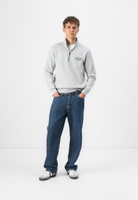 Grå zip-up sweatshirt med høj krave, parret med løse blå denim jeans og hvide sneakers med grå accenter. Enkel logodetalje på sweatshirt.