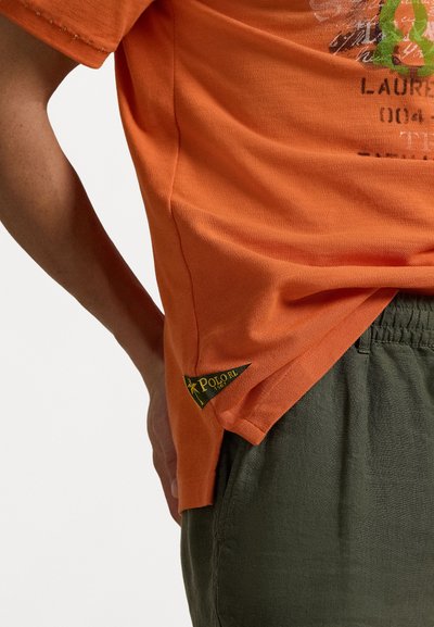 Oranje katoenen T-shirt met een gedrukt ontwerp op de voorkant en een logo label op de zoom. In combinatie met gestructureerde groene shorts.