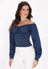 Top in denim con scollo off-shoulder, corpino arricciato, maniche lunghe e polsini arricciati. Presenta una consistenza morbida di colore blu scuro. Abbinato a pantaloni bianchi.