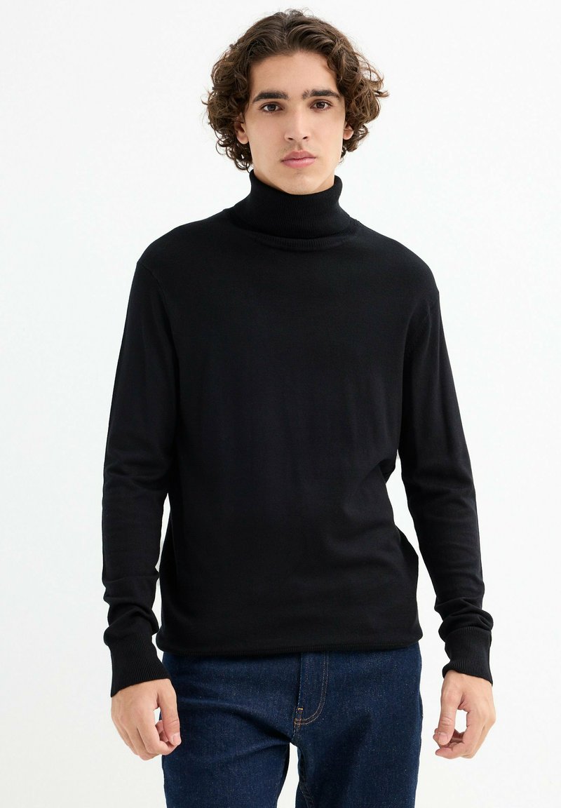 Jersey negro de cuello alto hecho de una tela suave, con mangas largas, puños de canalé y una textura lisa. Combinado con jeans azules.