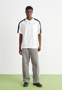 Camiseta atlética blanca con acentos negros en los hombros, de corte holgado, combinada con pantalones de pana grises y zapatos negros, de pie sobre una superficie verde.