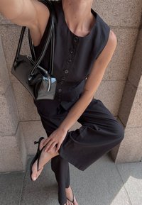 Zwarte mouwloze jumpsuit met een knoopsluiting, gecombineerd met een kleine zwarte lakleren handtas en zwarte slippers, tegen een gestructureerde achtergrond.