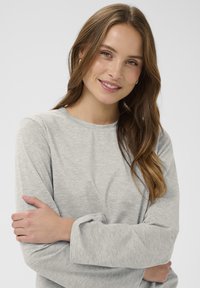 Smaidoša sieviete ar gariem brūniem matiem, tērpusies gaiši pelēkā garajā piedurkņu krekla, rokas mierīgi sakrustotas, pret vienkāršu baltu fona.