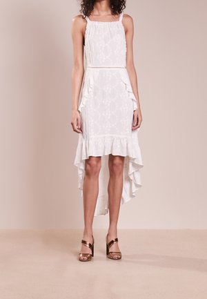 Mujer con vestido blanco sin mangas de corte asimétrico con borde fruncido y estampado floral, combinado con sandalias de tacón alto metálicas en color bronce.