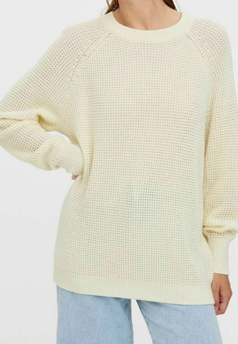 Jasnożółty, oversize sweter dzianinowy o teksturowanym, otwartym splotzie, z prążkowanym dekoltem i mankietami. Połączony z jasnoniebieskimi dżinsami.