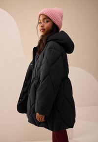 Veste matelassée noire avec capuche, taille élastique et fermeture par pression. Portée avec un bonnet en maille rose clair. Texture lisse, longueur jusqu'aux genoux.