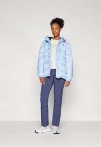 Donna in piedi che indossa una giacca imbottita azzurra chiaro, una maglietta con grafica bianca, pantaloni blu navy e sneakers bianche su uno sfondo bianco semplice.