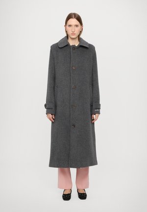 Lungo cappotto grigio in lana con colletto, chiusura frontale con bottoni, due tasche laterali e polsini arrotolati. Indossato sopra pantaloni rosa chiaro e scarpe nere.