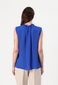 Mouwloze blauwe blouse met een geraffineerde halslijn. De gladde stof valt naar beneden in een ronde zoom. De achterkant toont subtiele plooien en een minimalistisch ontwerp.