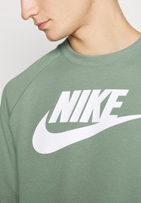 Zelená mikina vyrobená z měkké látky, která má velké bílé logo "NIKE" a swoosh. Design zahrnuje raglánové rukávy a kulatý výstřih.